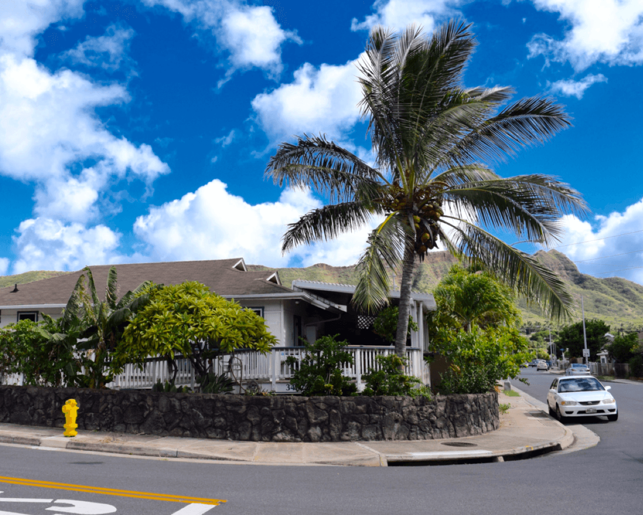 Hawaiian house with white porch on corner lot, tall palm tree, stone wall, yellow fire hydrant, mountains in background, blue sky.RetryClaude can make mistakes. Please double-check responses.